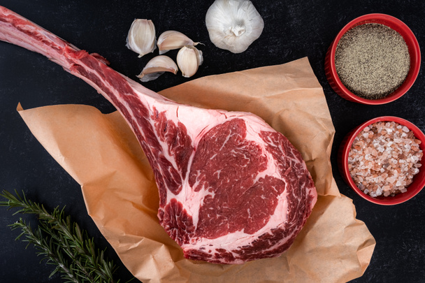 Frisch geschnittenes Tomahawk-Ribeye-Steak mit Metzgerpapier zum Würzen mit Salz und Pfeffer, Knoblauch und Rosmarin auf der Arbeitsplatte - Foto, Bild