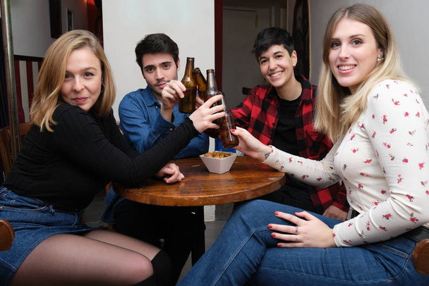 Vier lächelnde Freunde stoßen in einer Kneipe mit Bierflaschen an und schauen in die Kamera. Freundschaftskonzept. - Foto, Bild