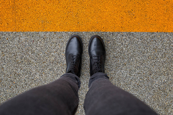 Μαύρα παπούτσια που στέκονται στο τσιμεντένιο δάπεδο με πορτοκαλί γραμμή. Πόδια που περπατούν σε εξωτερικούς χώρους. Νεαρός εγωιστής - Φωτογραφία, εικόνα
