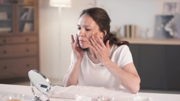 Medium close-up van geprepareerde en gepolijste vrouw van middelbare leeftijd met schoonheid routine verzorgen van haar huid het aanbrengen van zelfgemaakte exfoliërende scrub met koffiedik op het gezicht kijken naar spiegel - Video
