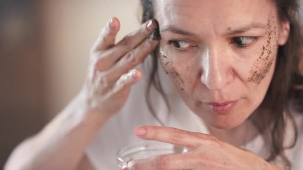 Close-up van geprepareerde gemengde ras vrouw in het midden van de 40 's toepassen van zelfgemaakte exfoliërende koffie scrub met verjongende effect masseren haar jukbeenderen kijken naar spiegel - Video