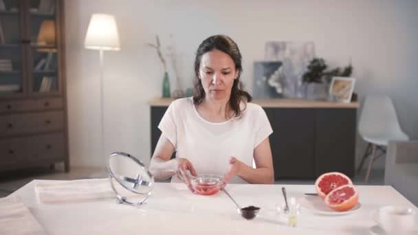 Medium shot van goed uitziende vrouw van middelbare leeftijd thuis zitten maken scrubben anti-age scrub door het toevoegen van koffiedik aan grapefruit pulp in kom - Video