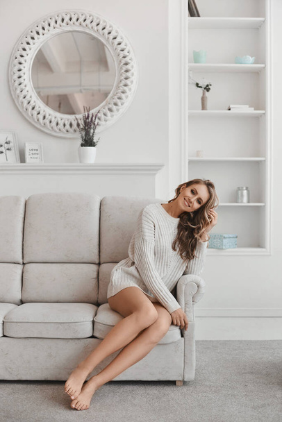 Young woman with long sexy legs and blond hair in white pullover posing on the sofa in a cozy apartment in the morning - Photo, image