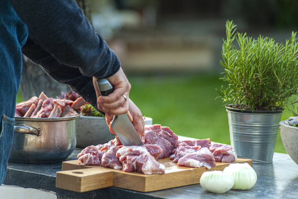 Preparation for BBQ: man's hand slicing meat and onion with big knife on wooden cut board, outdoors - Photo, Image