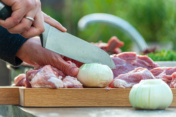 Preparation for BBQ: man's hand slicing onion and meat with big knife on wooden cut board, outdoors - Photo, Image