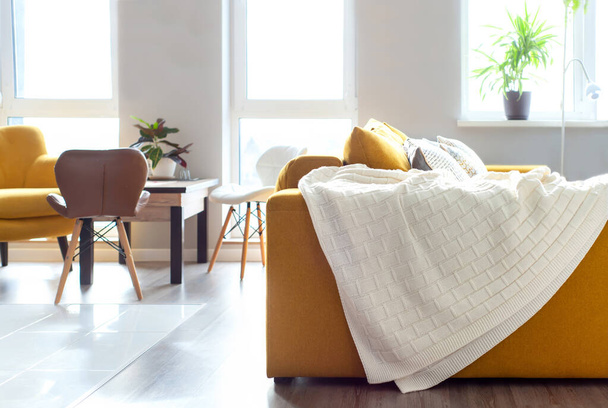Φωτεινό σαλόνι με κίτρινο καναπέ και λευκή πλεκτή κουβέρτα   - Φωτογραφία, εικόνα