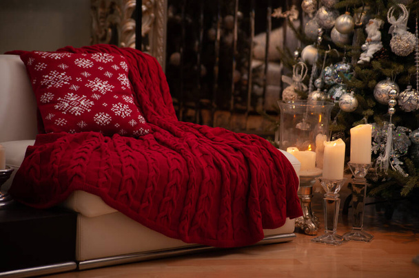 Nature morte de Noël avec magnifique couverture en tricot de câble rouge et coussin motif flocons de neige sur un canapé, grandes bougies sur un sol et arbre de Noël décoré sur le dos   - Photo, image