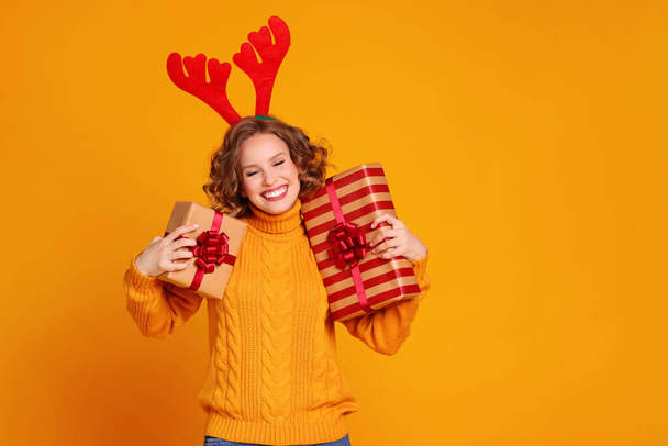 Šťastná mladá žena v pleteném svetru s červenými jeleny parohy ukazuje zabalené dárky a dívá se na fotoaparát s úsměvem na Štědrý den proti žlutému pozadí - Fotografie, Obrázek