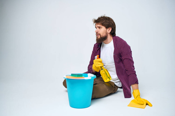 Een man ligt op de vloer met een emmer wasmiddel schoonmaak service lichte achtergrond - Foto, afbeelding