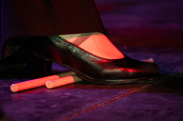 Bein einer Frau mit Hose, die einen schwarzen Flamenco-Tanzschuh trägt, unten ein paar hölzerne Schlagstöcke auf einer hölzernen Bühne, Licht auf der Bühne - Foto, Bild