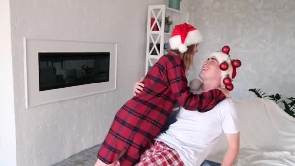 Laughing man and woman wearing santa hats and red pajamas fall together on the sofa cuddling. Young couple fooling around on christmas evening, slow motion - Footage, Video
