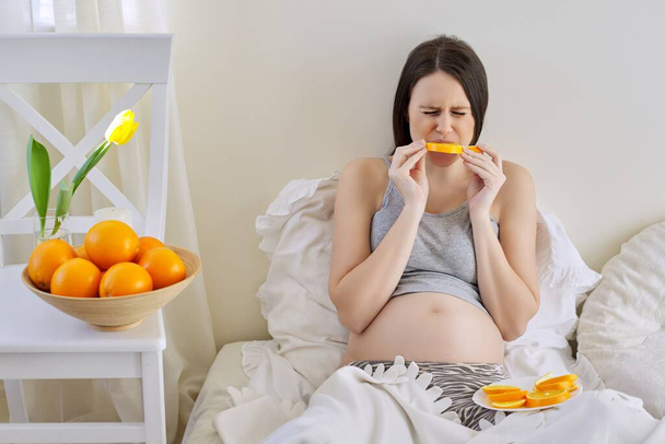 Young beautiful pregnant woman eating oranges sitting in bed at home - Foto, Bild