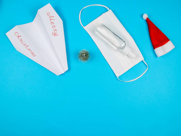 Avión, máscara, desinfectante, sombrero de santa y árbol de Navidad decorativo sobre un fondo azul con lugar para el texto de abajo, vista superior de cerca. - Foto, imagen