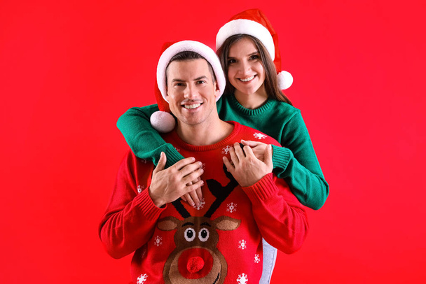 Beautiful happy couple in Santa hats and Christmas sweaters on red background - Fotografie, Obrázek