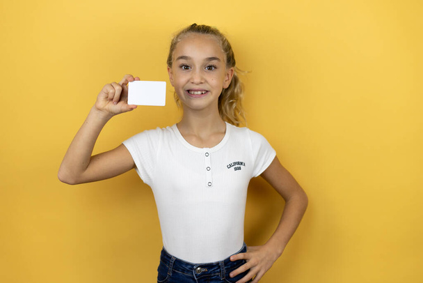 Young beautiful child girl standing over isolated yellow background smiling and holding white card - Fotografie, Obrázek