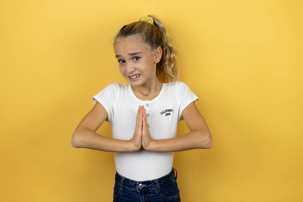 Young beautiful child girl standing over isolated yellow background begging and praying with hands together with hope expression on face very emotional and worried - Photo, Image