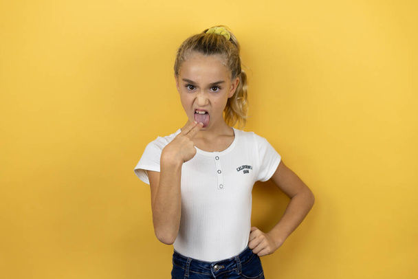 Junge schöne Kind Mädchen steht über isolierten gelben Hintergrund angewidert mit ihrer Hand im Mund - Foto, Bild