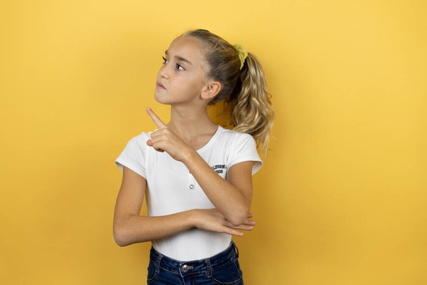 Young beautiful child girl standing over isolated yellow background smiling happy pointing with hand and finger to the side - Photo, Image
