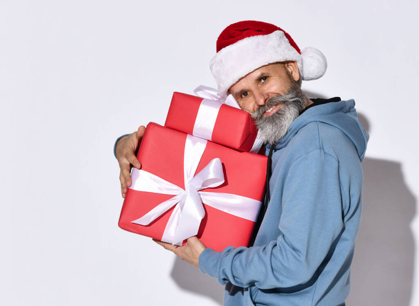 Senior bearded man points his finger at the gift boxes he holds on a white background. - Фото, изображение