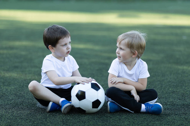 Životní styl dvou dětí a fotbalového míče, sedí na trávě, směje se a mluví. Koncepce dětství, sportu a komunikace předškolních chlapců. - Fotografie, Obrázek