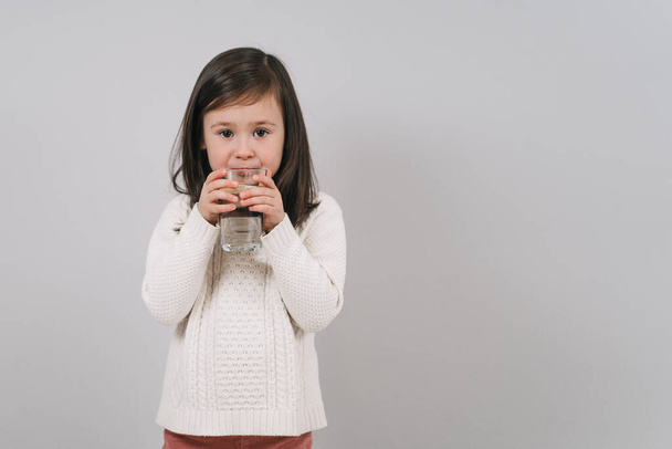 The child drinks water from a glass. A girl with dark hair holds a glass of water. The brunette leads a healthy lifestyle - 写真・画像