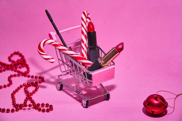 Shopping cart with red lipstick, candy cane, mascara. On a pink background. With a red Christmas ball and beads. Concept of Christmas shopping for beauty products.  - Photo, image