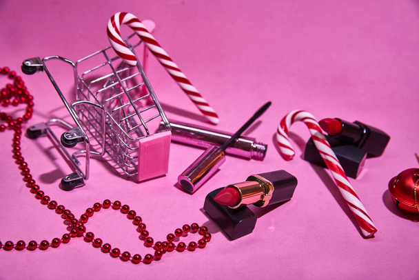 Shopping cart with red lipstick, candy cane, mascara. On a pink background. With a red Christmas ball and beads. Concept of Christmas shopping for beauty products.  - Foto, Imagem