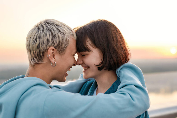 Close up of young loving lesbian couple hugging, laughing before kiss while admiring the sunrise together - Photo, Image
