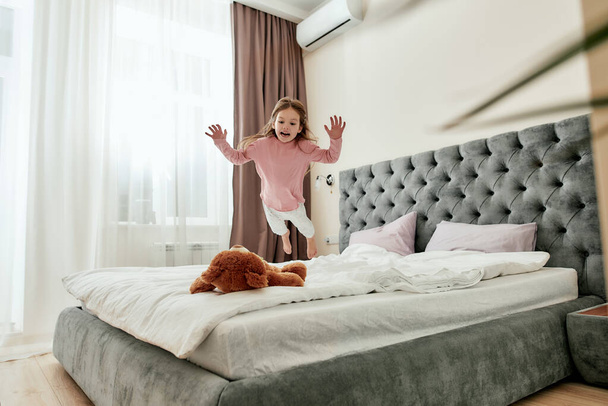 A cute small girl having fun and jumping down like a little tiger on her brown teddybear barefoot on a bed - Photo, image