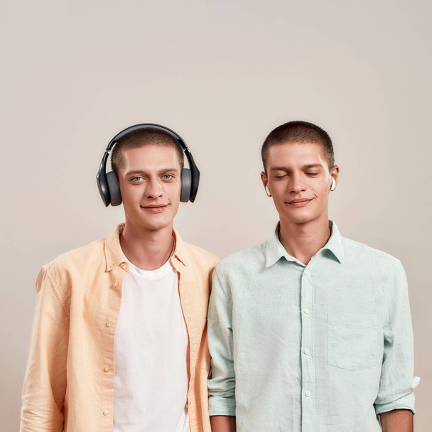 Two young caucasian twin brothers in casual wear listening music in headphones and wireless earphones, while standing isolated on beige background - Foto, immagini