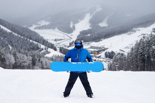 Ο τύπος κρατά στο χέρι μπλε snowboard το χειμώνα. Snowboarder σε μπλε σακάκι με χιονισμένα βουνά φόντο - Φωτογραφία, εικόνα