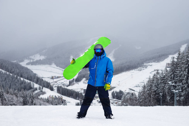 Парень держит на плече зеленый сноуборд зимой. Сноубордист в синей куртке на фоне снежных гор - Фото, изображение
