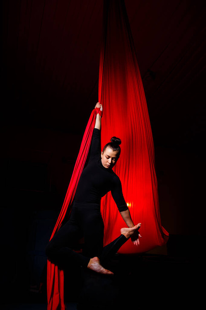 Ein Sportmädchen führt Turn- und Zirkusübungen auf roter Seide auf. Studioaufnahmen auf dunklem Hintergrund. Luftturnen auf Leinwand - Foto, Bild