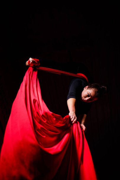 Ένα αθλητικό κορίτσι εκτελεί γυμναστικές ασκήσεις και ασκήσεις τσίρκου σε κόκκινο μετάξι. Γυρίσματα στούντιο σε σκοτεινό φόντο. Εναέρια γυμναστική σε καμβά - Φωτογραφία, εικόνα