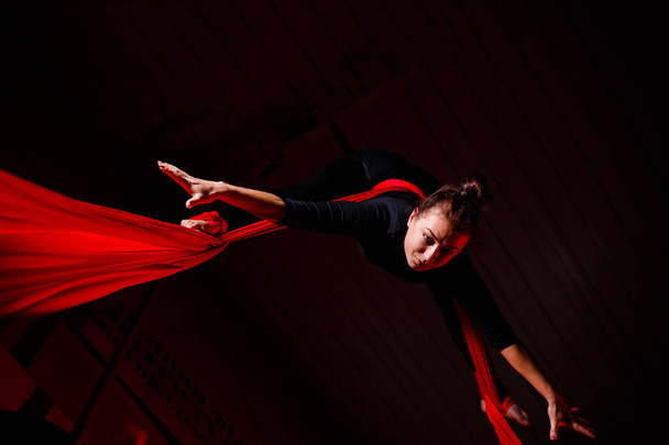 Sportovní dívka provádí gymnastická a cirkusová cvičení na červeném hedvábí. Studio natáčí na tmavém pozadí. Letecká gymnastika na plátně - Fotografie, Obrázek
