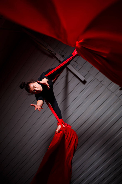 A sports girl performs gymnastic and circus exercises on red silk. Studio shooting on a dark background. Aerial gymnastics on canvas - Фото, изображение