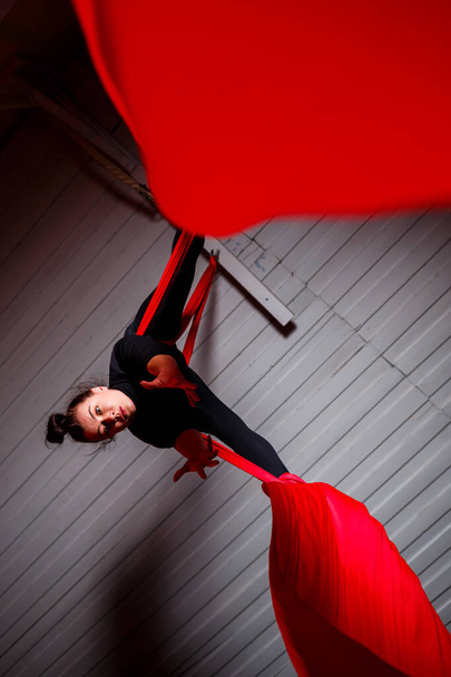 Urheilutyttö suorittaa voimistelu- ja sirkusharjoituksia punaisella silkillä. Studio ampuu pimeällä taustalla. Ilmavoimistelu kankaalle - Valokuva, kuva