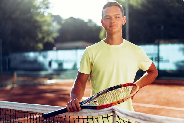 Young man teenager with tennis racket standing near net on clay court - Fotografie, Obrázek