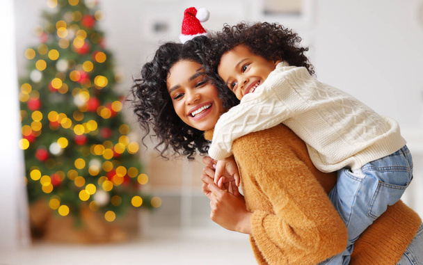 Side view of happy ethnic woman giving piggyback ride to boy and smiling while having fun at home during Christmas celebratio - Foto, afbeelding