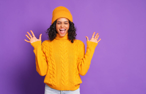 Entzückte junge Frau in gestricktem gelben Pullover und Hut gestikuliert und schreit mit geschlossenen Augen, während sie vor lila Hintergrund Spaß hat - Foto, Bild