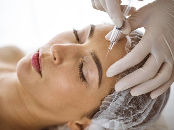 Beautician doing beauty procedure with syringe to face of young brunette woman in sunny spa center. Cosmetic medicine and surgery, beauty injections - Foto, Imagen
