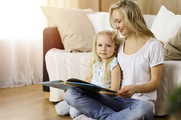Happy family. Blonde young mother reading a book to her cute daughter while sitting at wooden floor in sunny room. Motherhood concept - Foto, afbeelding