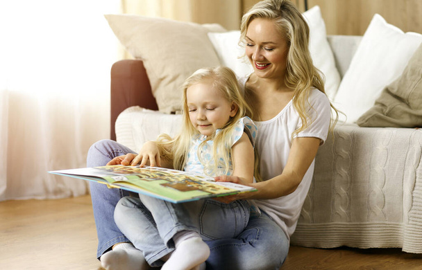 Happy family. Blonde young mother reading a book to her cute daughter while sitting at wooden floor in sunny room. Motherhood concept - Φωτογραφία, εικόνα