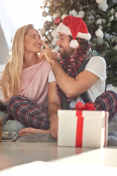 beardy guy touching nose of his girlfriend with santa hat on his head, smiling. christmastime - Фото, изображение