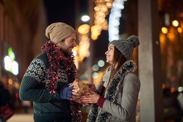 A boyfriend giving a Christmas present to his girlfriend while walking the city together on a beautiful holiday night. Christmas, relationship, love - Zdjęcie, obraz