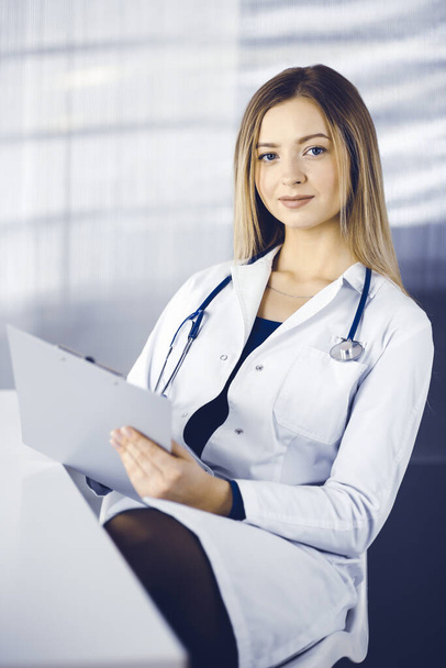 Young optimistic woman-doctor is using a clipboard, while sitting at the desk in her cabinet in a clinic. Portrait of friendly female physician woman with a stethoscope. Perfect medical service in a - Фото, изображение