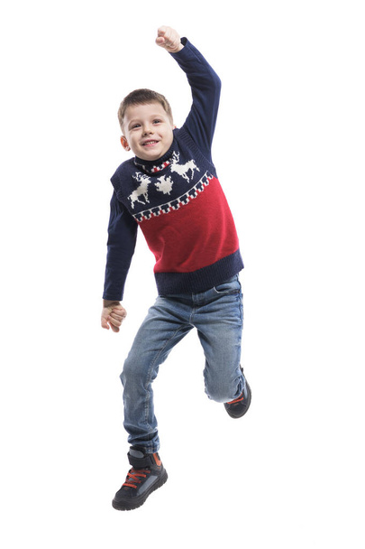 Радостный маленький ребенок прыгает взволнованный в рождественский свитер глядя вверх с сжатыми кулаками. Полный портрет тела изолирован на белом фоне.  - Фото, изображение