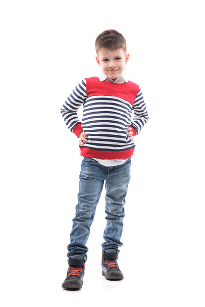 Élégant petit garçon heureux posant en pull et jeans avec les mains sur les hanches. Portrait intégral isolé sur fond blanc.  - Photo, image