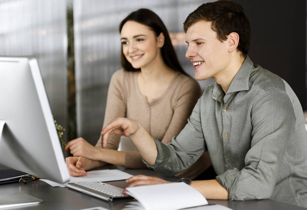 Friendly young businessman and programmer in a green shirt is working on computer, while sitting together at the desk with a female colleague in a modern office. Focus on man. Concept of successful - Фото, изображение