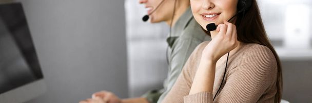 Two young people in headsets are talking to the clients, while sitting at the desk in an office. Focus on woman. Call center operators at work - 写真・画像